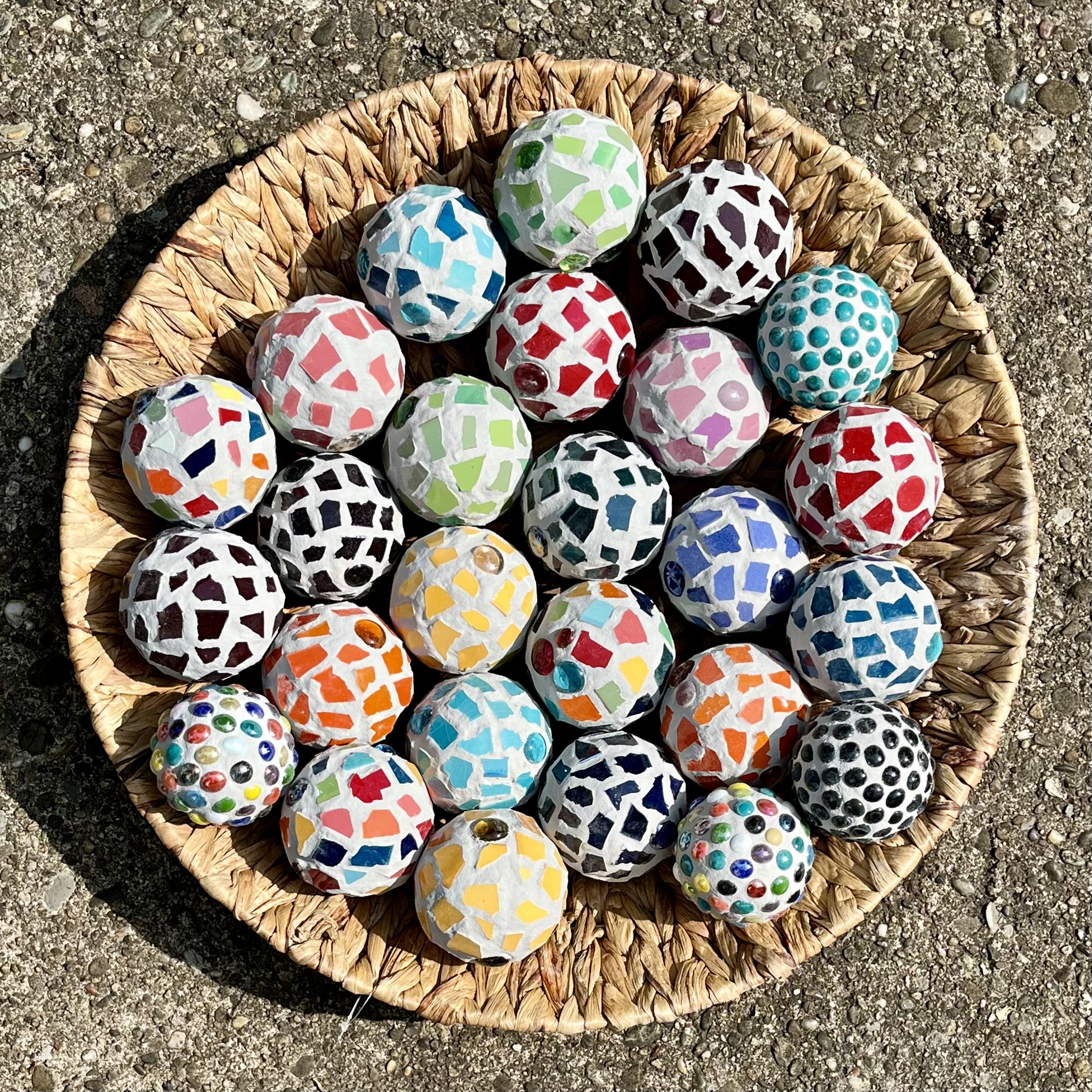 Bunte Mosaikbälle in einem geflochtenen Korb, verschiedene Farben und Muster, auf einem grauen Untergrund.