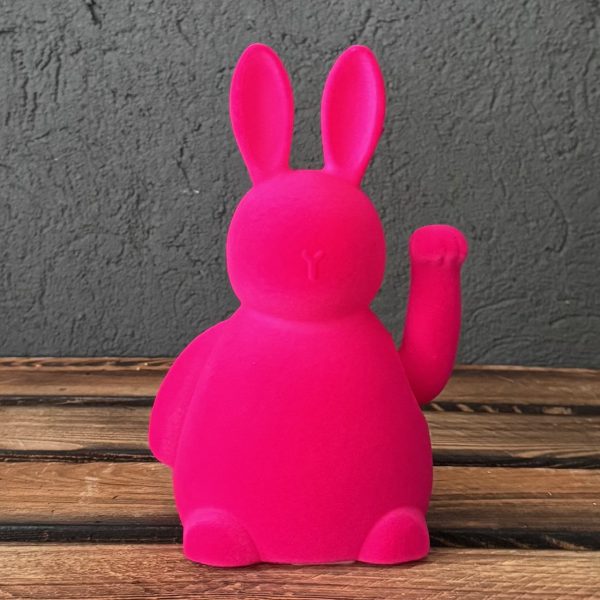Rosa, niedlicher Hase mit langen Ohren, einer Hand, die winkt, auf einem Holztisch vor grauem Hintergrund.