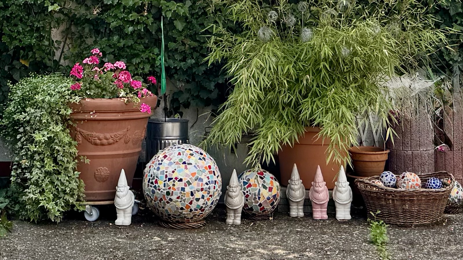 Bunte Gartenlandschaft mit Tontöpfen, rosa Blumen, einem Mosaikball und mehreren Gartenzwerge.